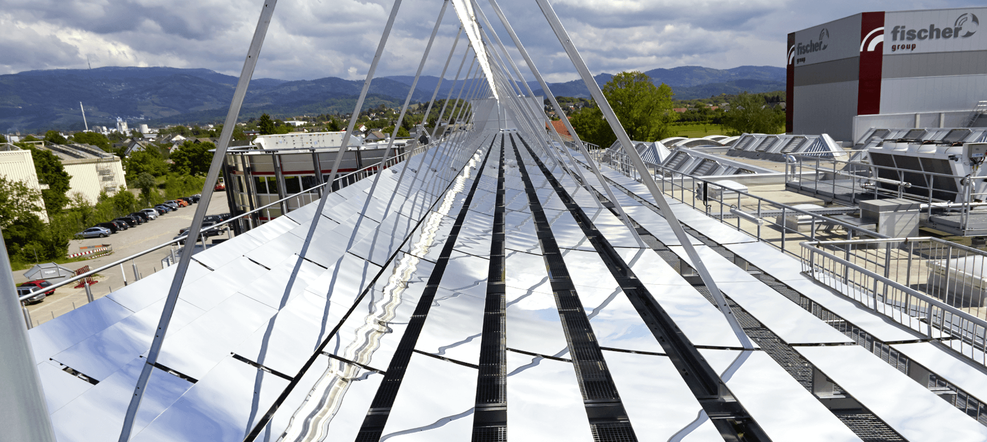 Eine stark spiegelnde Solaranlage installiert auf einer der Fabrikhallen von fischer in Achern. Entlang der Solarmodule ist der Blick gerichtet auf die Höhen des Schwarzwaldes. Links und rechts der Solaranlage sind weitere Fabrikhallen der Firma fischer zu sehen.